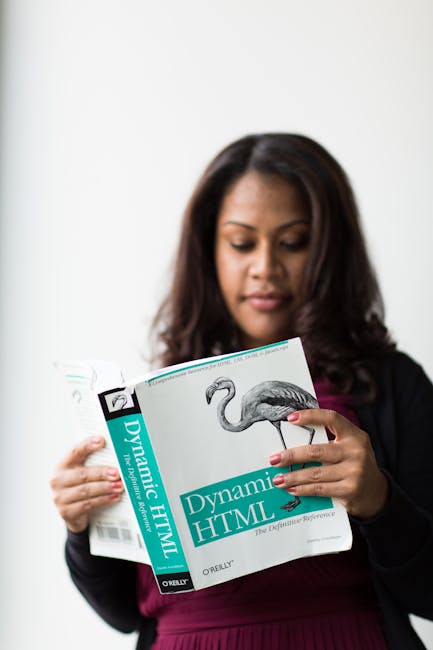 A woman with dark, wavy hair is standing indoors, holding and reading a large book titled 'Dynamic HTML' by Oreilly, which features an illustrated flamingo on the cover. She is dressed in a dark blaze
