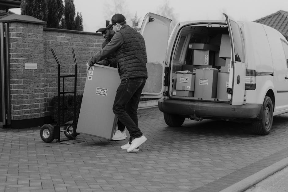 A man wearing a blue beanie, a navy and red windbreaker, black trousers, and white sneakers with black and brown accents sits on the edge of an open van's loading area. The interior of the van contain